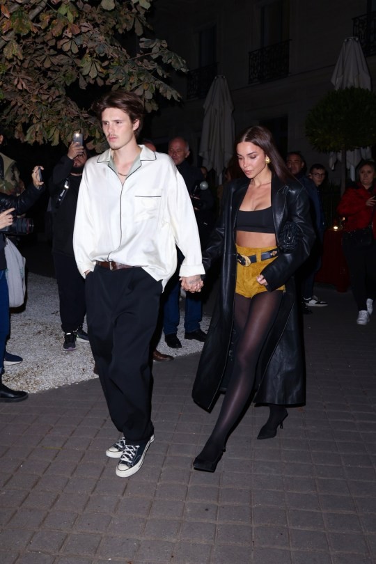 Cruz Beckham and Jackie Apostel hold hands while walking in Paris. Cruz is wearing black trousers and a white shirt. Jackie is wearing yellow shorts with a black top, leather coat, and tights