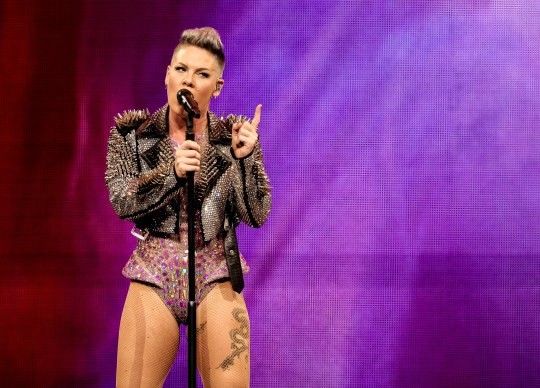 Pink sings into a microphone onstage while wearing a spiked black leather jacket over an embellished pink leotard