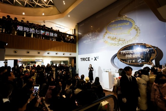 a crowded promotional event featuring a speaker in front of a large screen displaying jewelry and signage