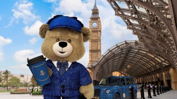 a dressed teddy bear holding a coffee cup near a notable clock tower