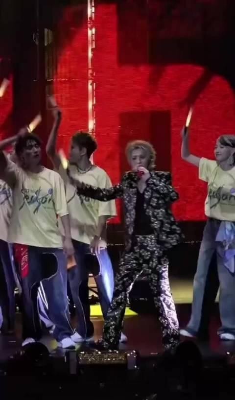 舞台表演中，主角身穿黑色花纹套装，手持麦克风，周围多名伴舞穿黄色T恤，背景为红色LED屏幕。