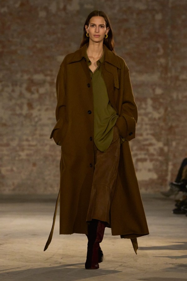 female model walking on a runway wearing a long brown coat and green shirt