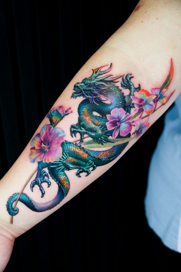 龙与花纹身 – 一幅传统亚洲风格的龙纹，以绿色和蓝色为主调，与粉紫色花朵交织在前臂上。构图融合了动态动感与繁复的花卉细节。