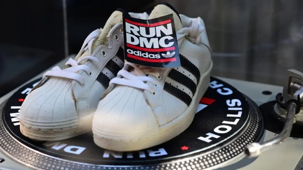 一双破旧的白色阿迪达斯Superstar运动鞋，带有黑色条纹，摆放在转盘上，上面有RUN DMC和Adidas Originals的标签。庆祝这一嘻哈乐队成立40周年的弹出式艺术装置展的一部分。