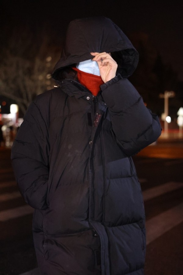 照片中人身穿黑色羽绒服及红色高领毛衣，并戴着口罩和帽子，包裹得相当严实。