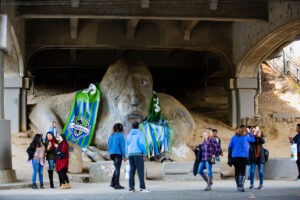 A large troll statue under a concrete bridge is draped with colorful sports jerseys. Several people stand around, some posing for photos and others walking or talking nearby.