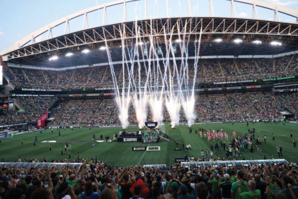A large stadium filled with cheering fans as fireworks shoot into the sky from a stage on the soccer field during a post-match celebration. Photographers and players gather near the stage, and the stands are packed with people.
