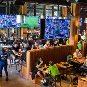 People sit at tables and booths in a lively sports bar, watching baseball on large TVs hanging above the bar area. Some wear team shirts, while others eat, drink, and talk. The atmosphere is busy and energetic.