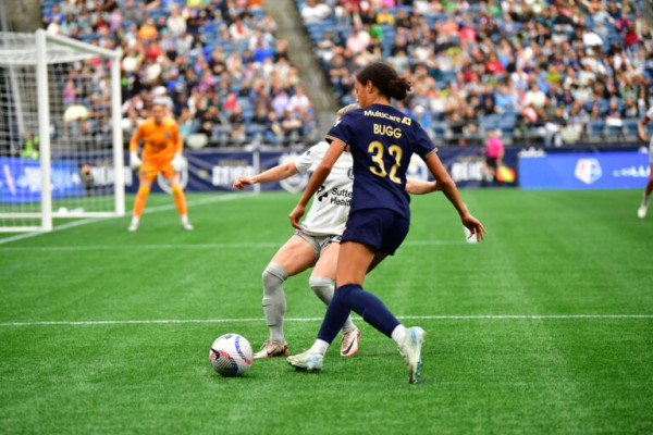 A soccer player in a navy blue jersey with "32 BUGG" on the back is about to kick the ball, while a defender in a white kit blocks her. A goalkeeper and a crowd of spectators are in the background.